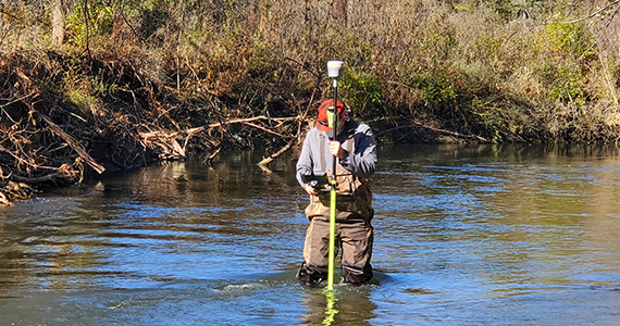 Staff member wearing waders and standing in the middle of a river with survey equipment