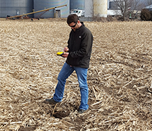 Staff member using a GPS unit, standing over a small sink hole in a farm field.